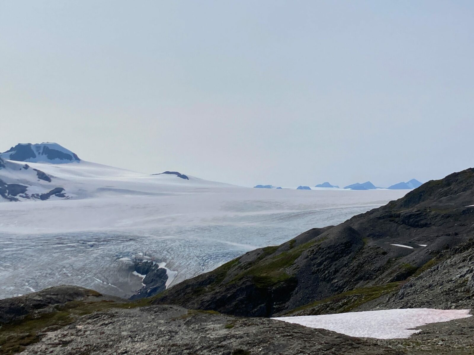 In the foreground are rocky mountains with a glacier next to them. In the distance there are more mountains sticking up from a massive icefield behind the glacier