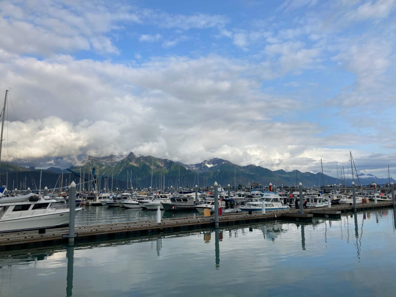 The boat harbor in Seward Alaska. There are many wooden docks with pleasure boats and fishing boats tied up. There are mountains across the fjord from the harbor that are partially obscured by clouds on an otherwise sunny day