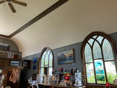 The interior of a former church that is now an art gallery in Seward, Alaska. There are two tall windows with stained glass art and many smaller art pieces on shelves below.