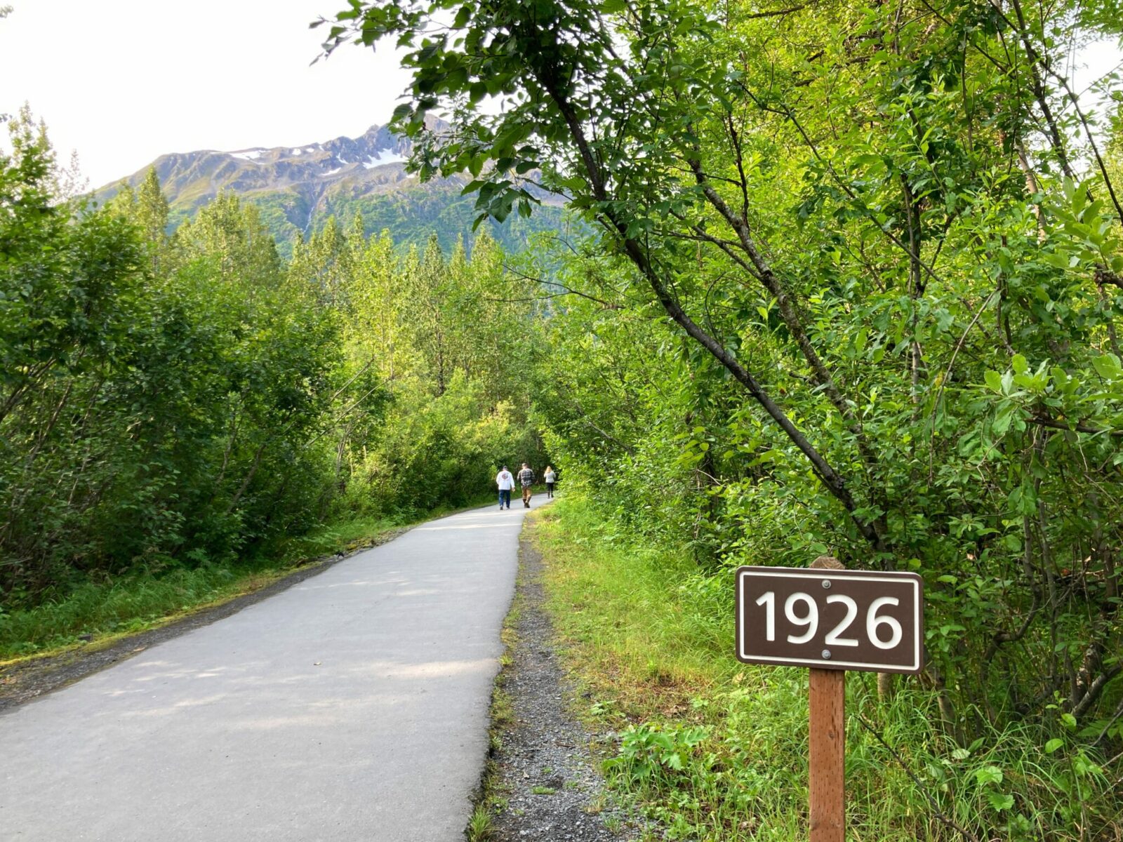 A paved trail through the forest. A wooden sign that says "1926" is on the side of the trail, marking the location of the toe of Exit Glacier in 1926.