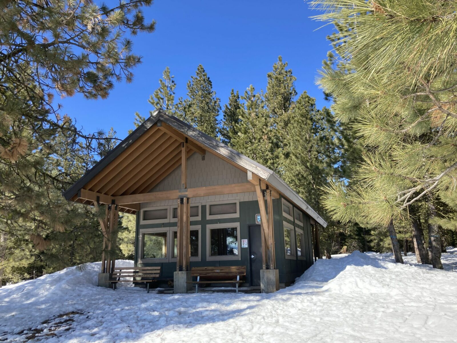 The Puffer Butte warming shelter in fields spring state park. The shelter is a wooden structure with big windows and a steeply pitched roof. There are two benches next to the front door. It is surrounded by snow and there are ponderosa pine trees surrounding it