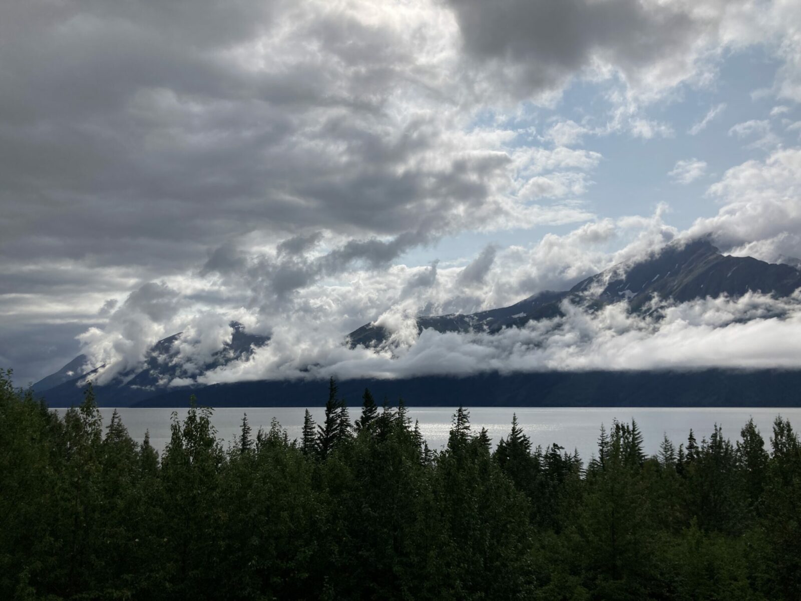 On a dark day, the sun tries to poke through white and gray clouds around mountains. There is water and forest in the foreground.