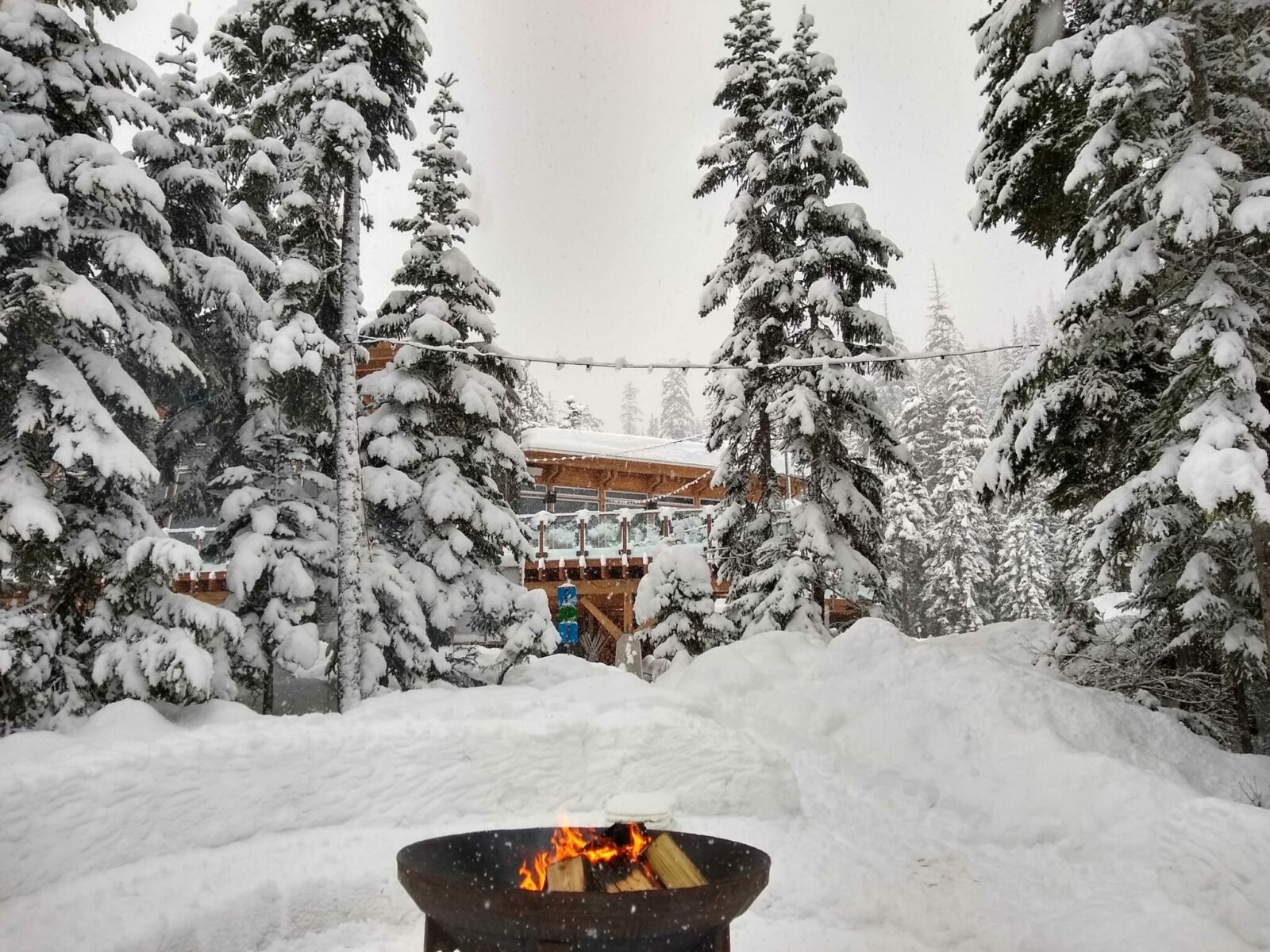 A fire pit with a wood fire in the snow surrounded by snowy forest. In the background is a log lodge