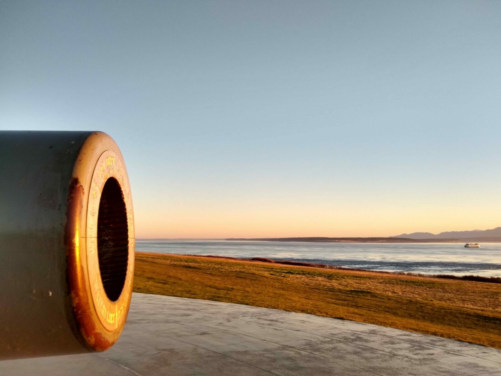 An old cannon at fort casey state park on whidbey island at sunset. There is a field and distant mountains and a ferry in the background