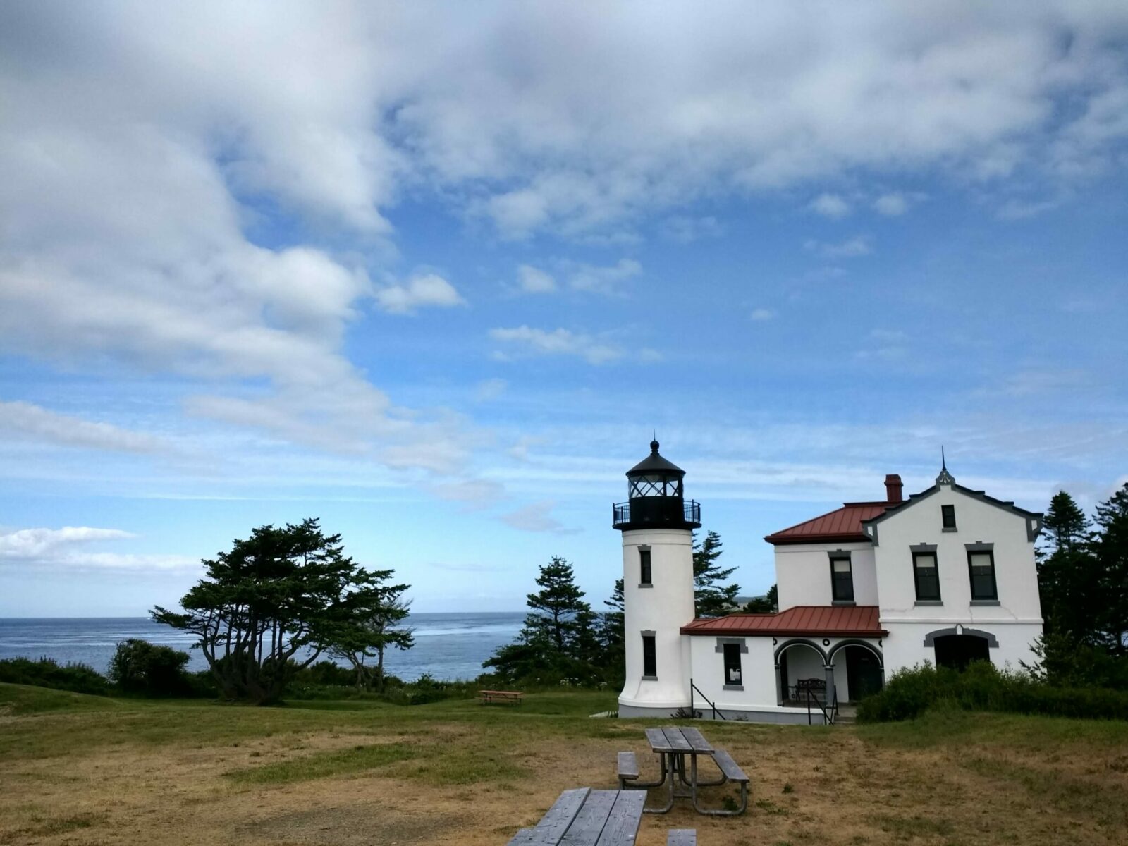 Admiralty head lighthouse on whidbey island. The lighthouse is in a field next to the rocky shore. It is white with a red roof and black trim. The actual light housing is black. There are two picnic tables in the foreground and some evergreen trees around the lighthouse. The partly cloudy sky and ocean are in the background