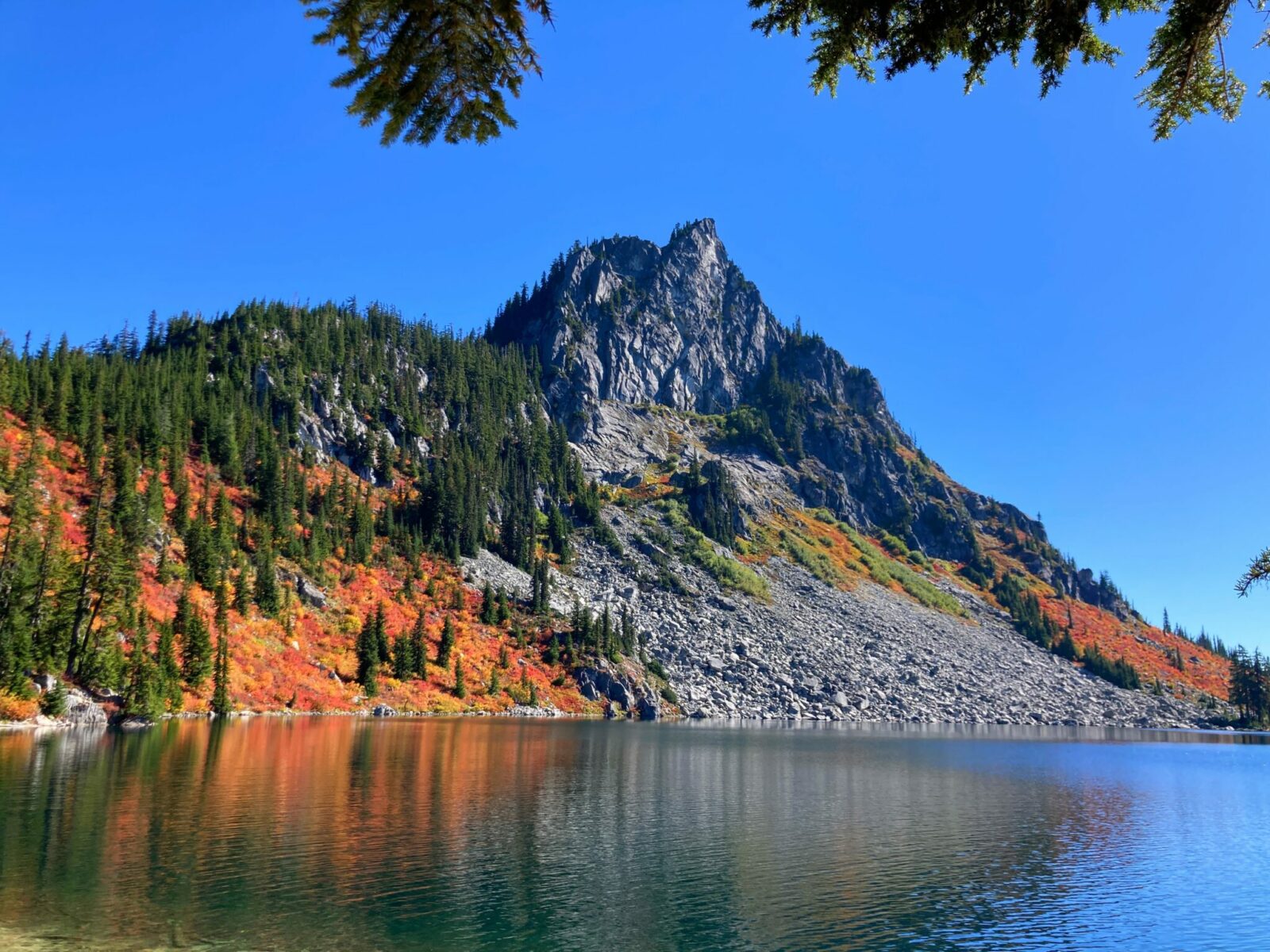 Calm, alpine Lake Valhalla with a rocky mountain on the opposite side of the lake. There are evergreen trees, gray rocks and bright orange and red berry bushes in fall color around the mountain across the lake