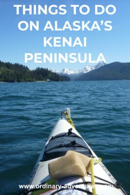 The front of a white kayak from the perspective of the person paddling it. In the distance are high snow capped mountains and evergreen forested hillsides. Text reads: things to do on alaska's kenai peninsula