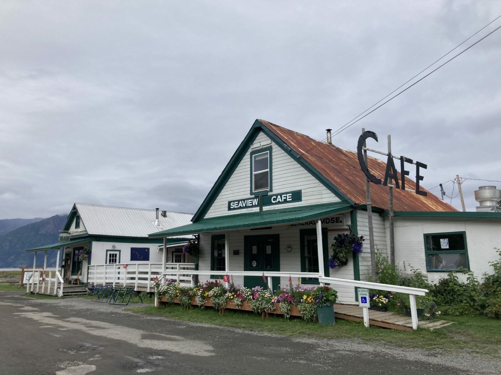 A historic white building with green trim and a metal roof with a large sign saying "cafe". There are some flowers on the porch and it is on a dirt road