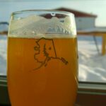 a beer in a glass next to a window with snow outside at a brewery in alaska. The glass has the outine of the shape of the state of alaska