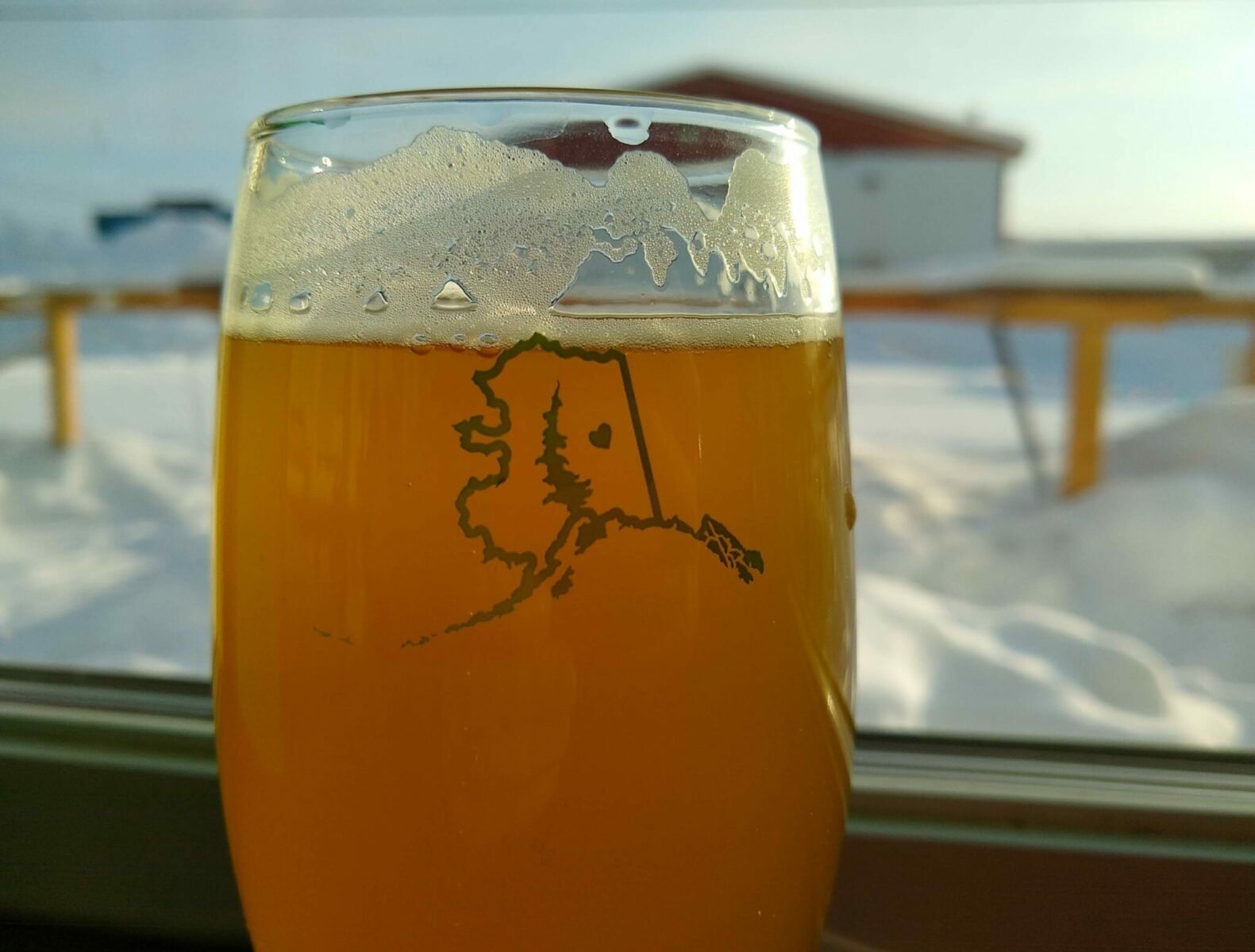 a beer in a glass next to a window with snow outside at a brewery in alaska. The glass has the outine of the shape of the state of alaska