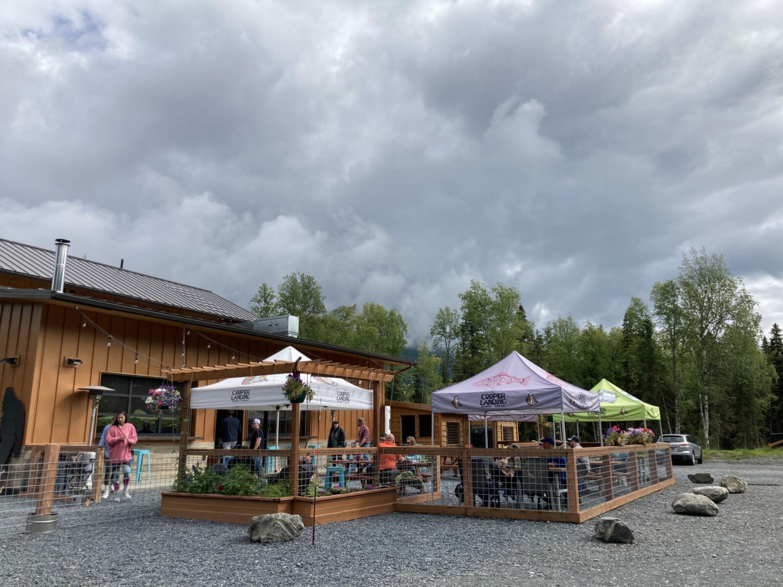 A fence with tables and tents over the tables at one of the breweries in Alaska. There is a large brown building and trees around a gravel parking lot next to the tents