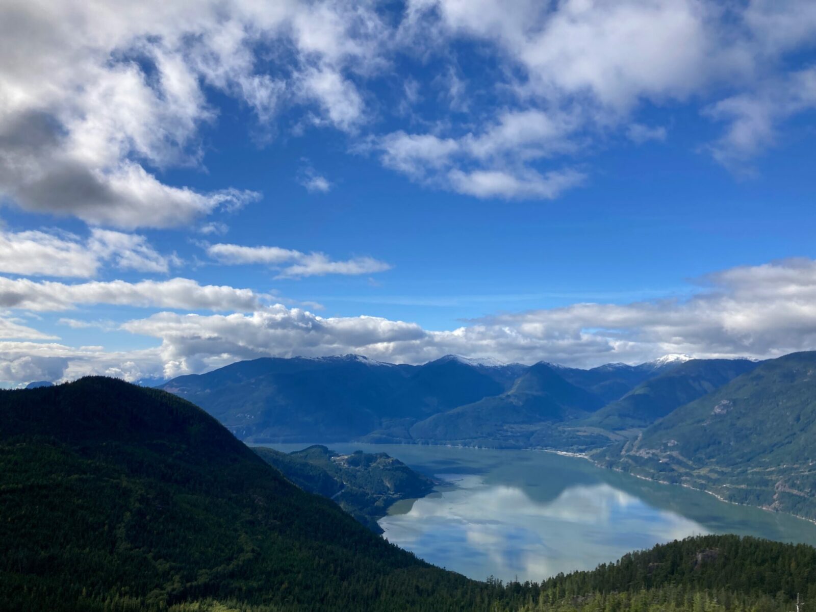 View from the top of the Sea to Sky Gondola on the Vancouver to Whistler drive. It's a partly cloudy day and there are near and distant mountains with a fjord and forests below