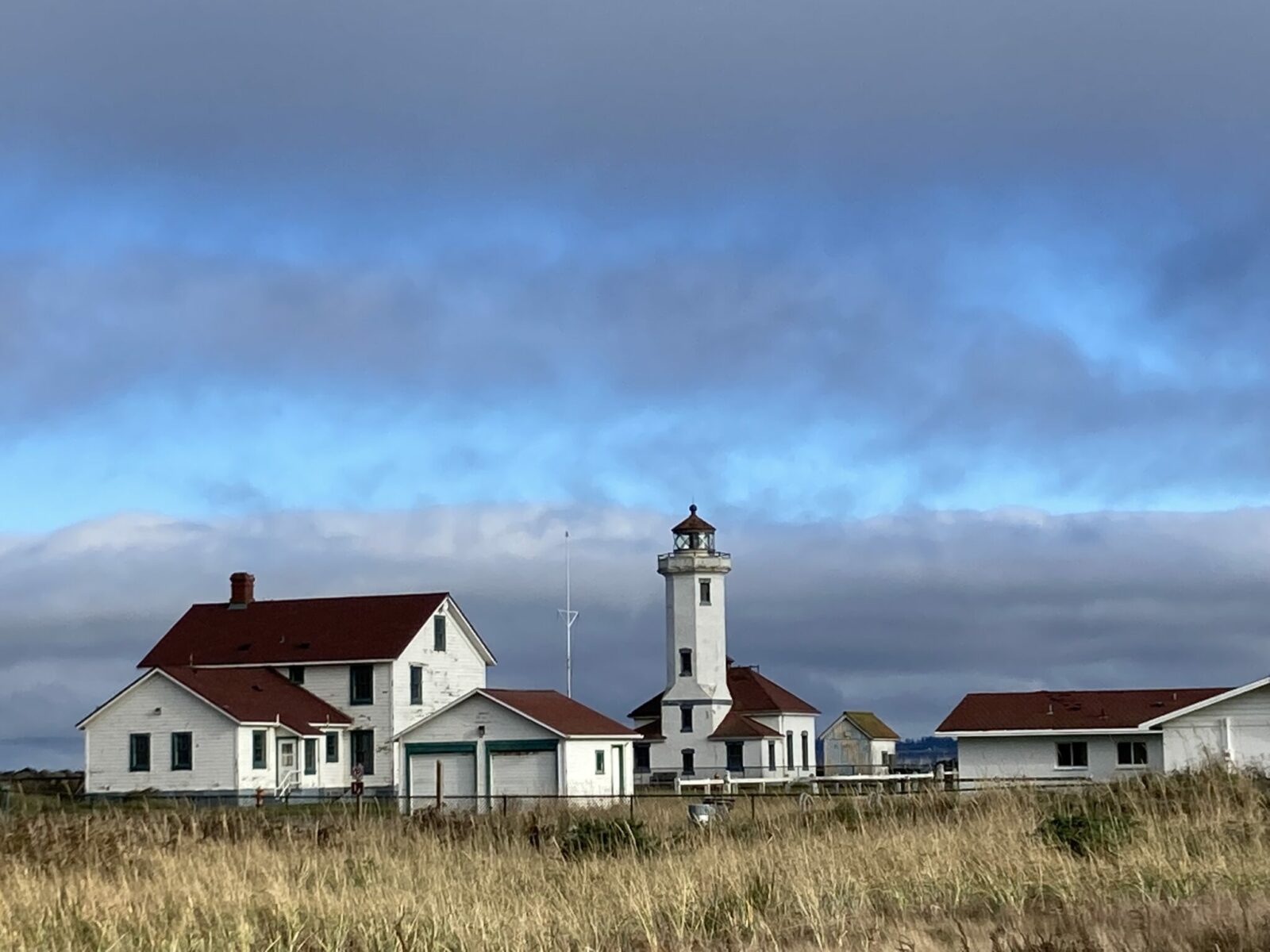 The Point Wilson Lighthouse complex. There is the lighthouse, a quarters for people to live, a garage and a modern building. All the buildings are white with red roofs. There is grass and a fence in the foreground.