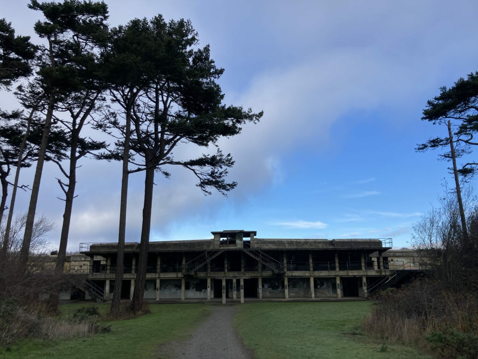 The ruins of a concrete military building. It is two stories and doesn't have doors. There is grass and trees surrounding it.