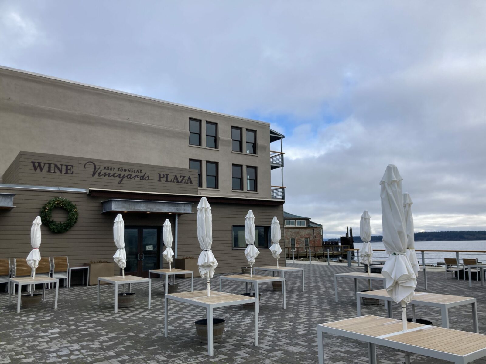 A stone plaza with tables and umbrellas folded up on a winter day. The building next to the plaza has a sign that reads Port Townsend Vineyards wine Plaza.