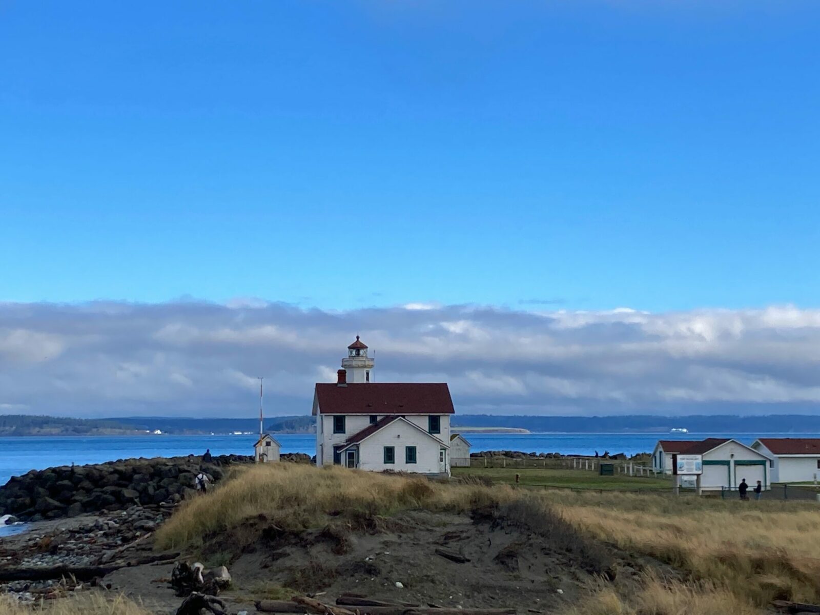 A lighthouse in a field next to a beach in Port Townsend. The lighthouse is a white building with a red roof. It is a partly sunny and partly cloudy day.