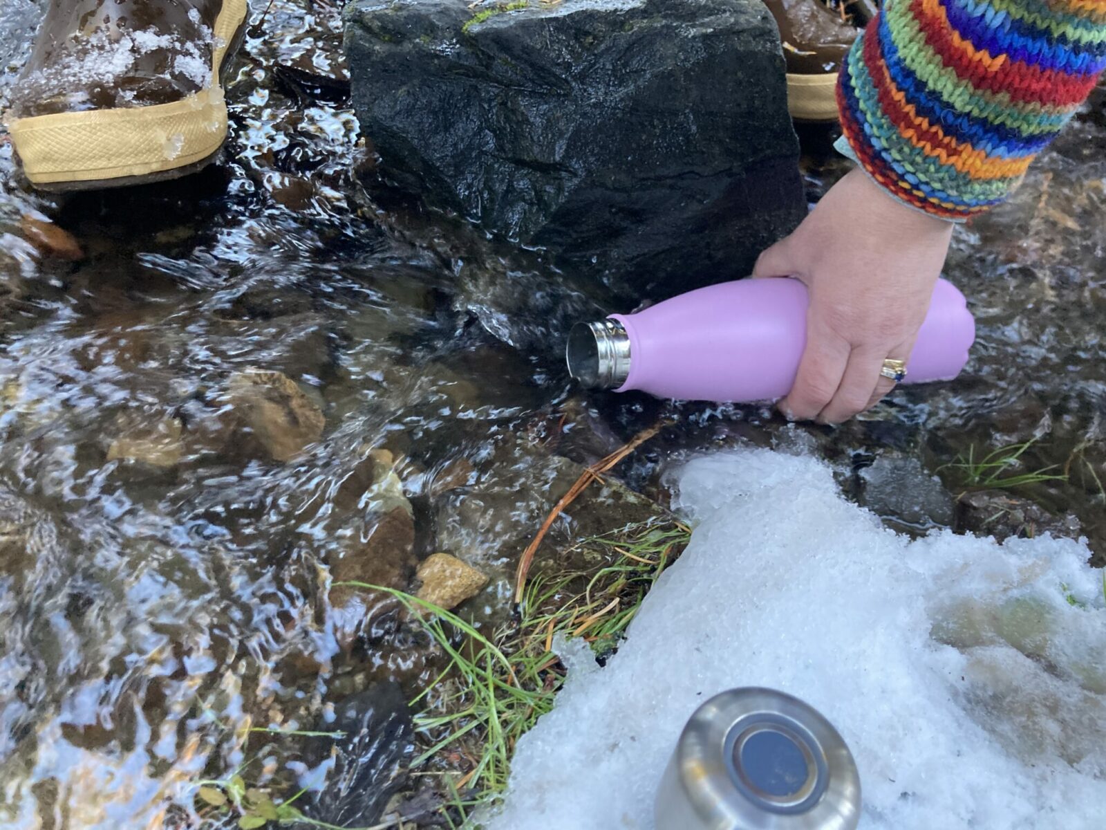 CrazyCap 2, a metal water bottle with a UV water purifier in the lid, being filled in a small stream with snow surrounding it. The bottle is in the water and the cap is sitting next to it. The water is flowing over rocks and a person's hand is holding the bottle in the water