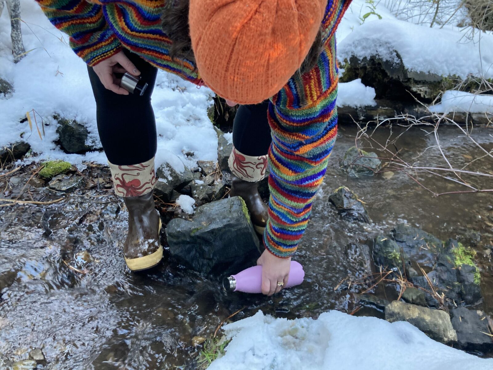 CrazyCap 2, a metal water bottle with a UV water purifier in the lid, being filled in a small stream with snow surrounding it. The bottle is in the water. The water is flowing over rocks and a person's hand is holding the bottle in the water. The person is wearing a colorful sweater, orange hat, black leggings and brown rubber boots with octopuses on them