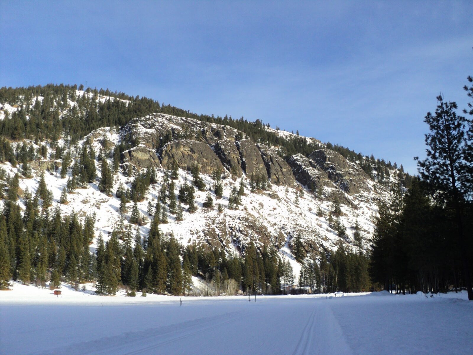 The cross country ski trails in Winthrop and the Methow Valley. A groomed ski track in shadow at the edge of a pine forest with a rocky tree and snow covered hill in the background