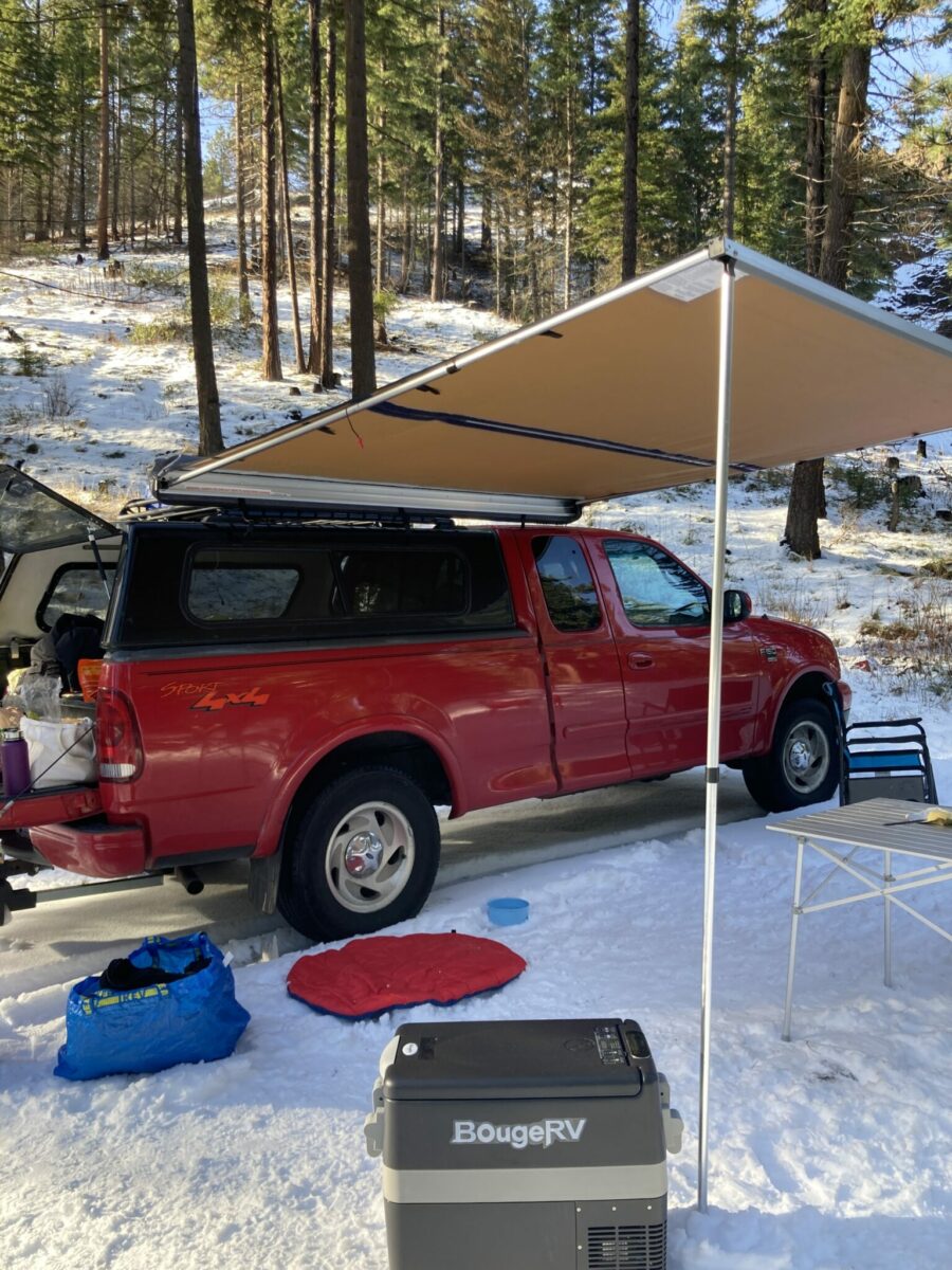 An electric cooler at a camp site. It is a Bouge RV brand electric cooler sitting in the snow next to a red pick up truck with an awning, camp chairs and a table.