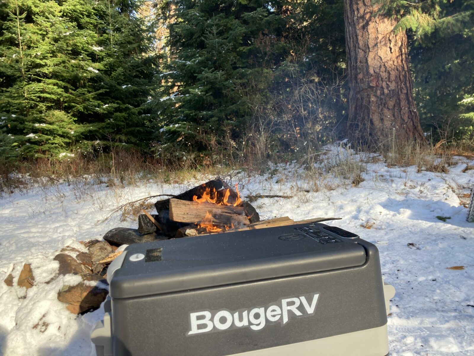 A Bouge RV brand electric cooler sitting outside next to a campfire on a snowy day