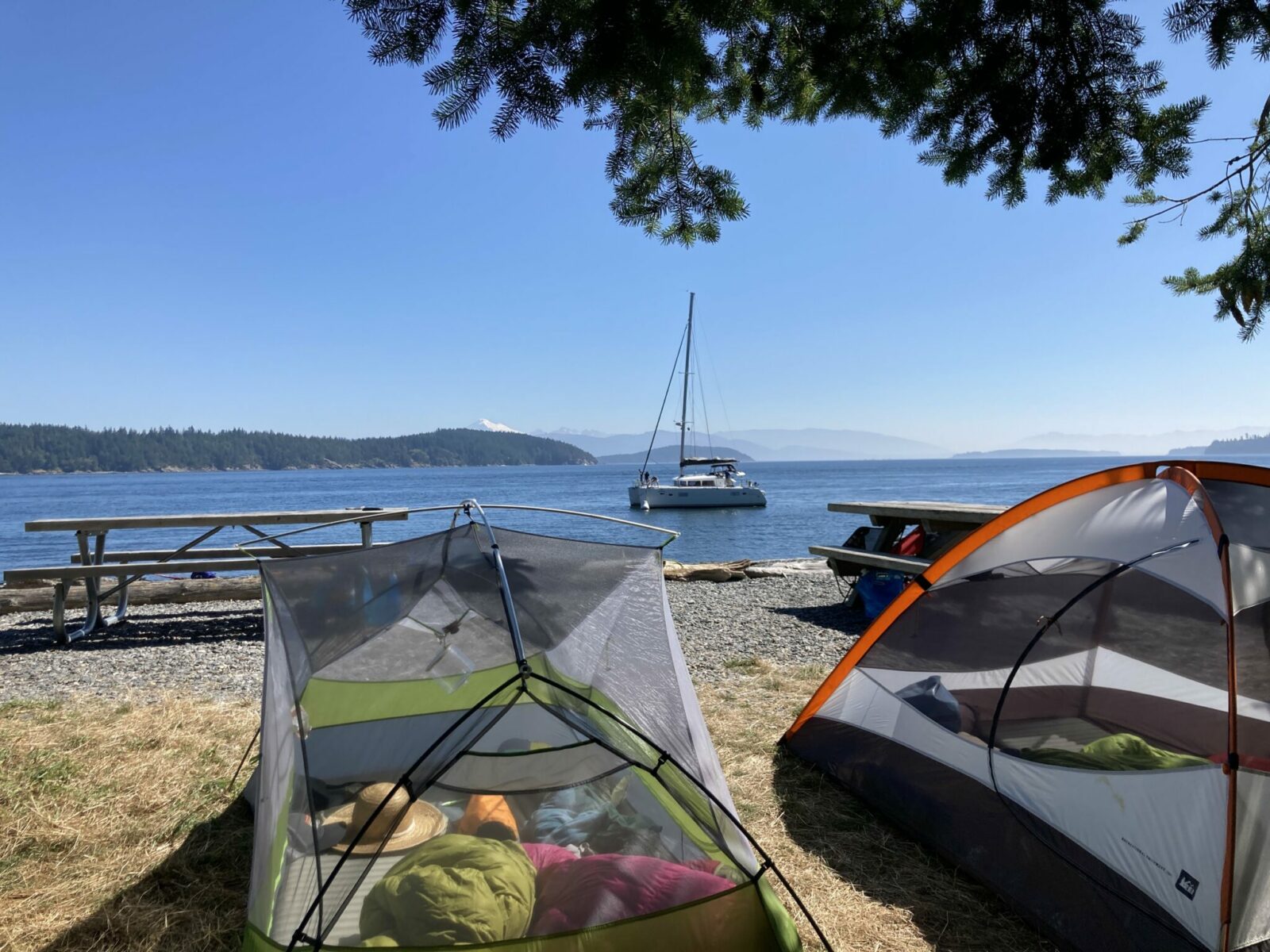 Two tents next to picnic tables at Pelican Beach on Cypress Island. The tents have gear and clothing in them and are in the mid day sun. There is a sailboat anchored just off the beach and there are forested islands and mountains in the distance.