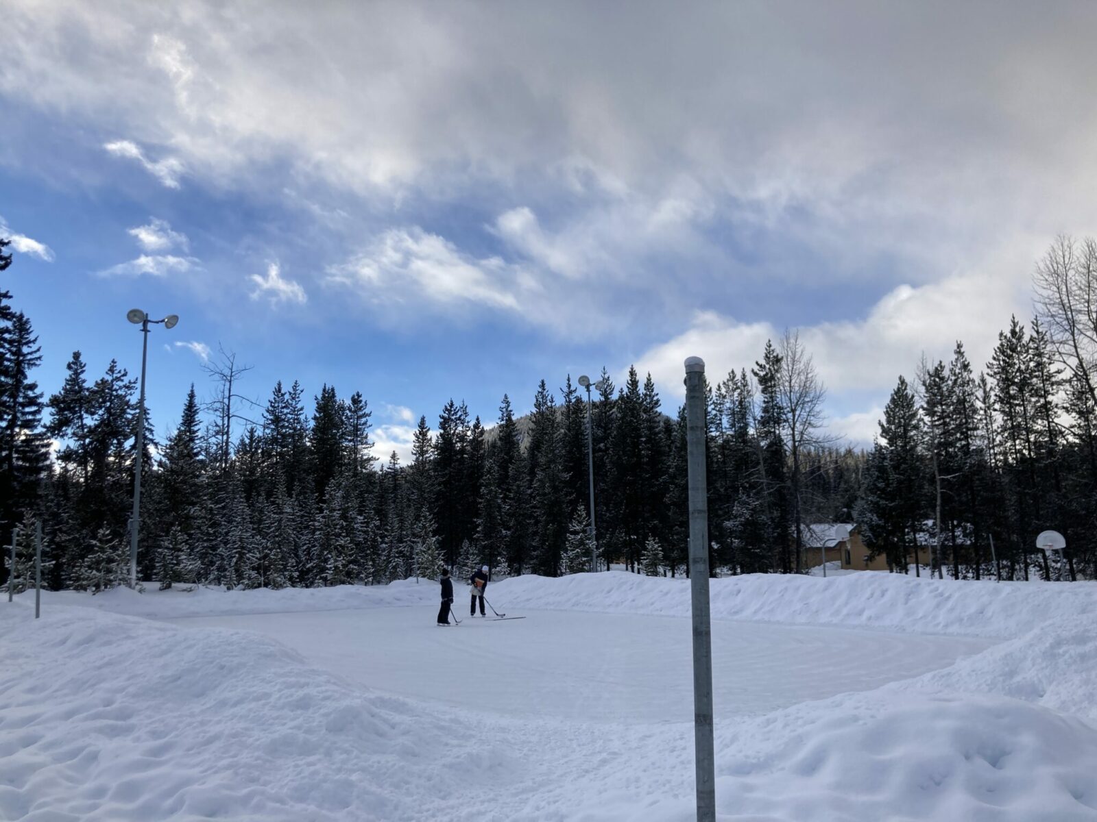 Two skaters on an outdoor ice rink in Manning Park. There are trees surrounding the rink and lights mounted on poles.