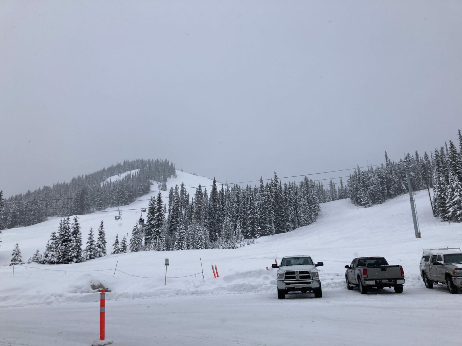 The base of a ski area with some snowy trees and a chairlift and a few parked pick up trucks in the foreground.