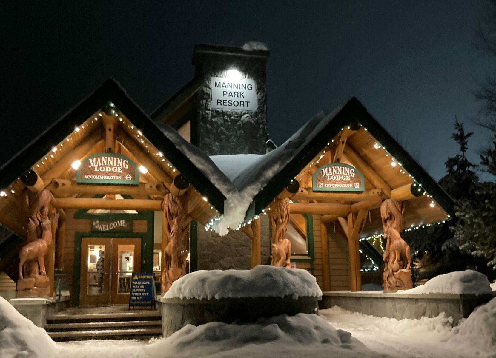 A wooden and stone lodge in winter. There is snow surrounding the lodge and the sign reads Manning Park Resort. There are white lights lighting up the entrance