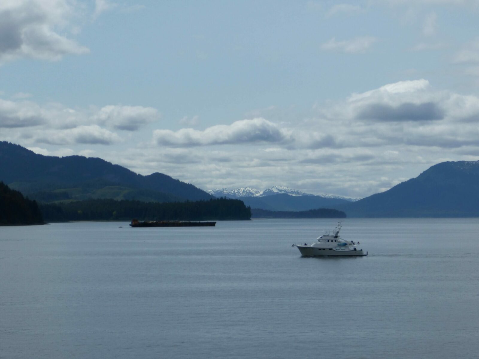 A boat and a barge in the water surrounded by forested hills and high mountains on a cloudy day