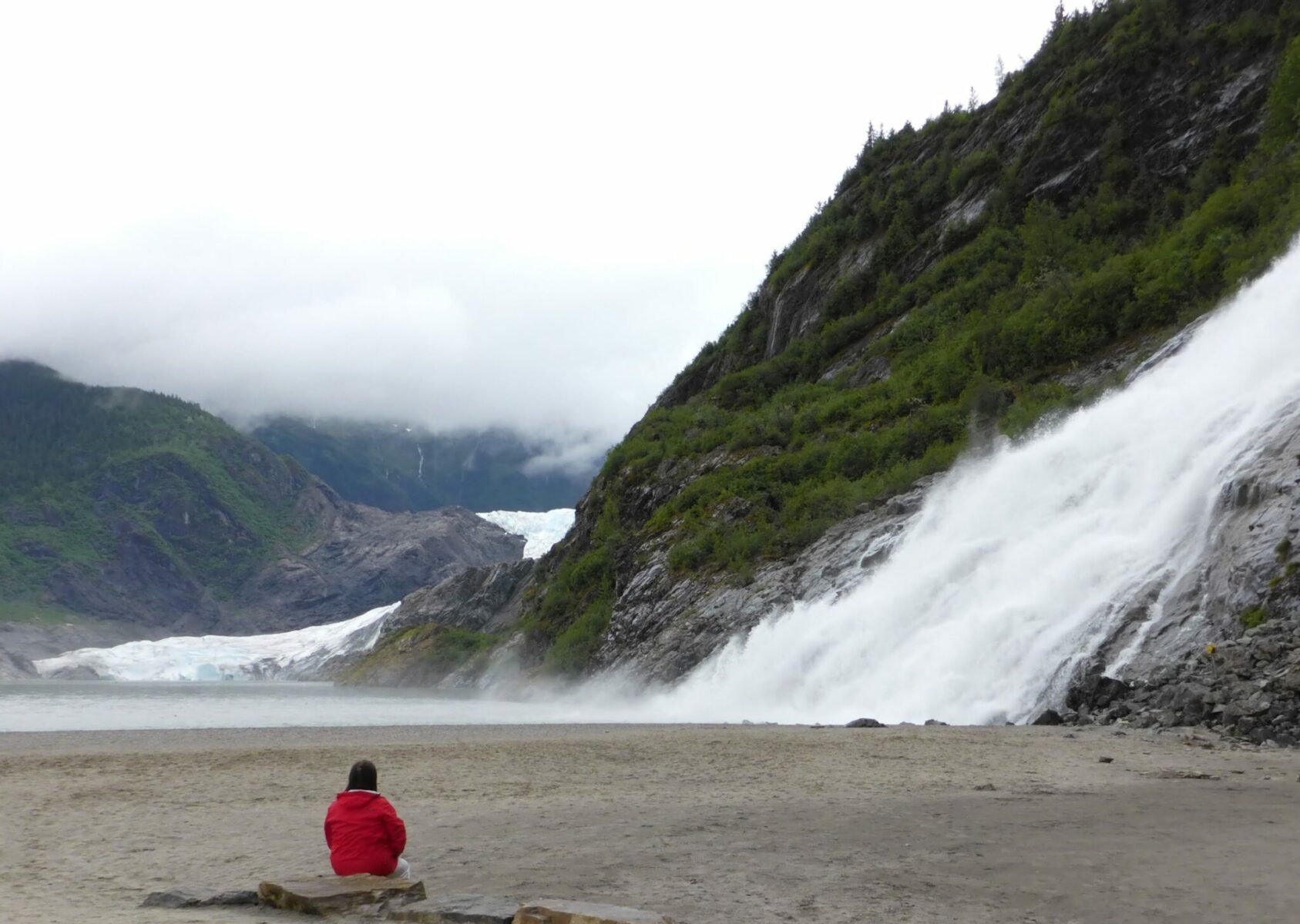 A waterfall coming down a rocky and brushy hill near a glacier. There is a large sandy area below the waterfall and a person in a red jacket sitting on a rock looking at the waterfall