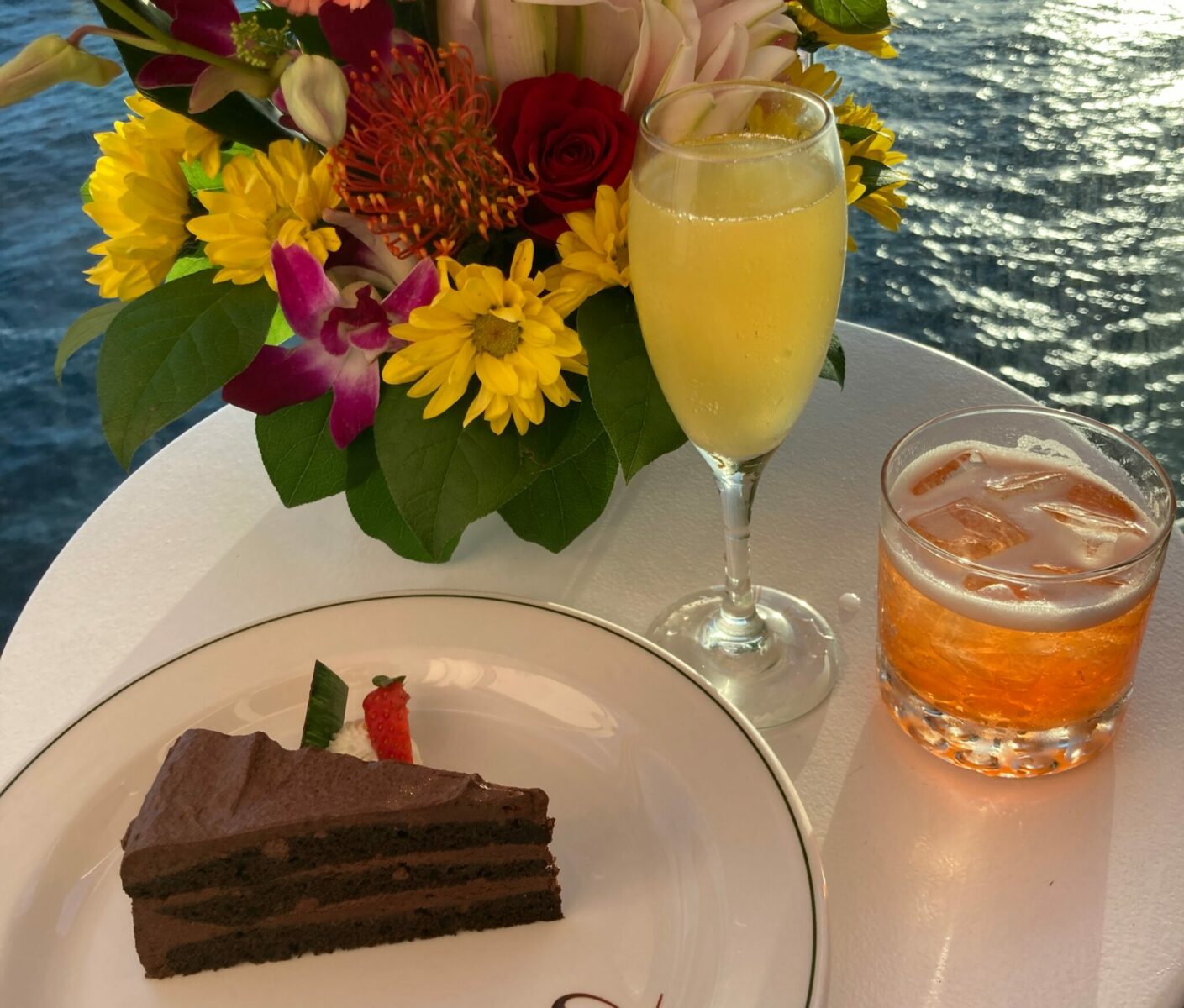 A small white table on the balcony of a cruise ship at sunset. There is a bouquet of flowers as well as two cocktails and a piece of chocolate cake.