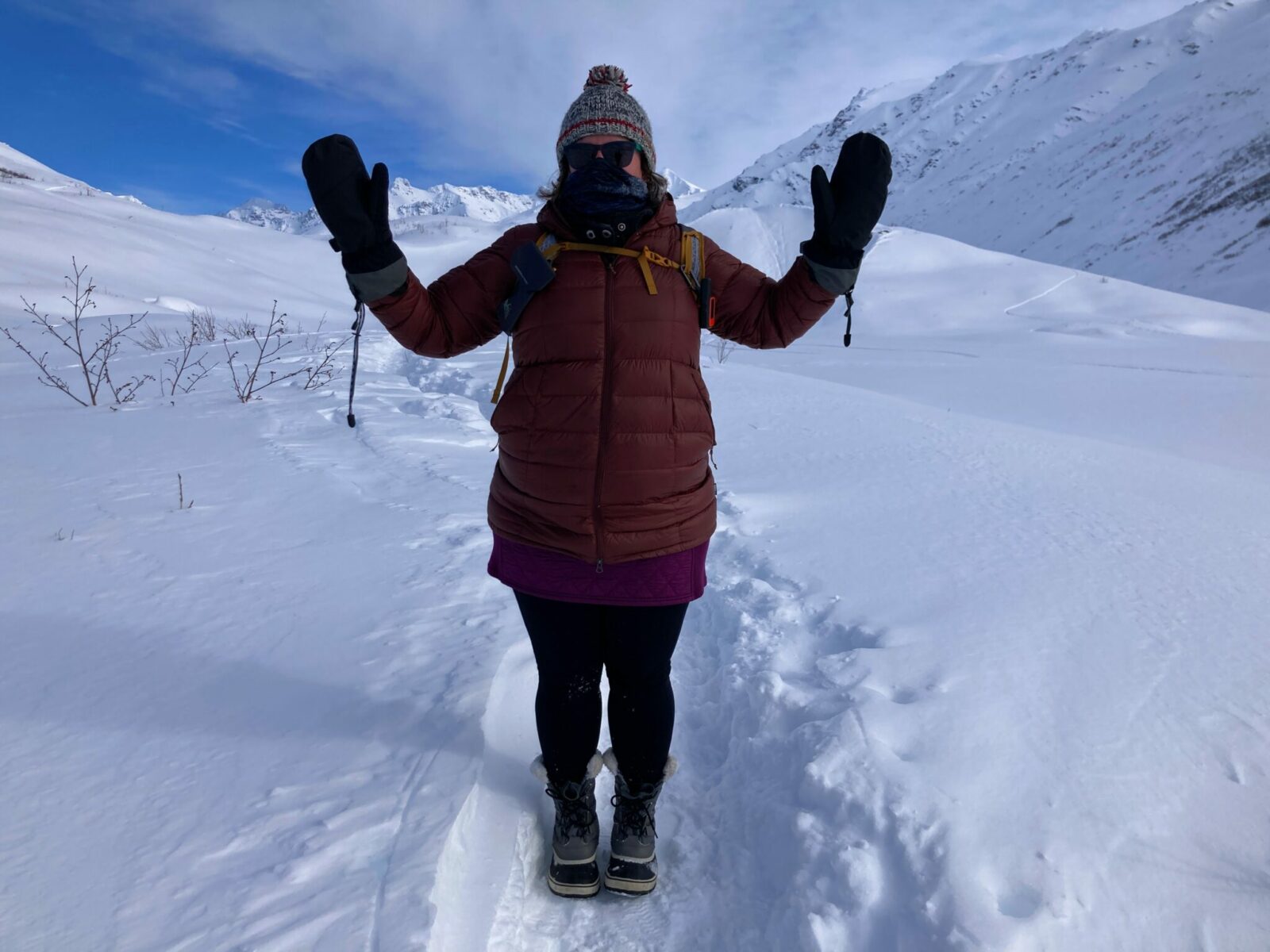 a hiker wearing a gray wool hat, a brown down coat, black leggings, snow boots, black mittens, a buff and sunglasses along with a backpack on a snowy trail in the mountains.