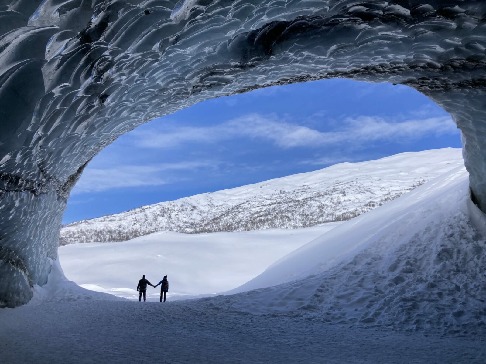 two people stand holding hands at the entrance to the ice cave at Castner Glacier. The photo is taken from inside the cave looking out. There is ice below and above and all around and a snowy landscape can be seen outside the cave