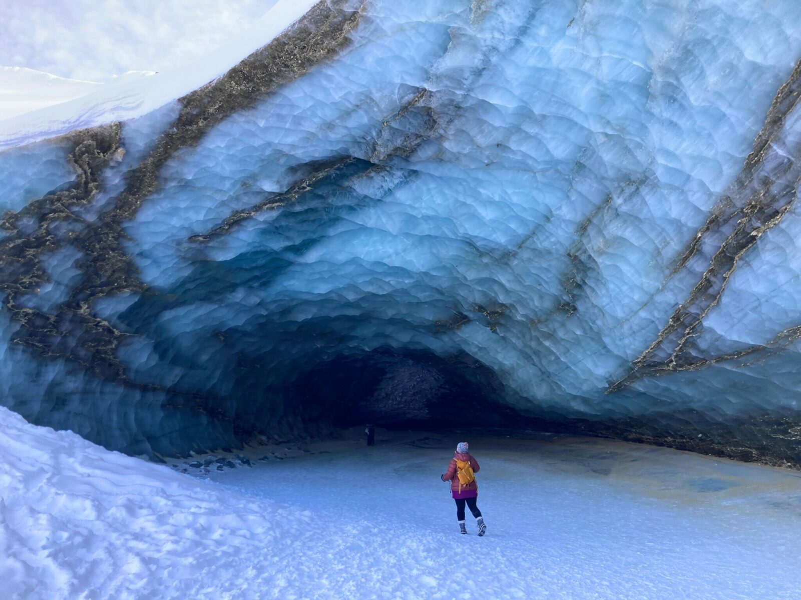 A person at the edge of an ice cave that is streaked with blue and brown rocks