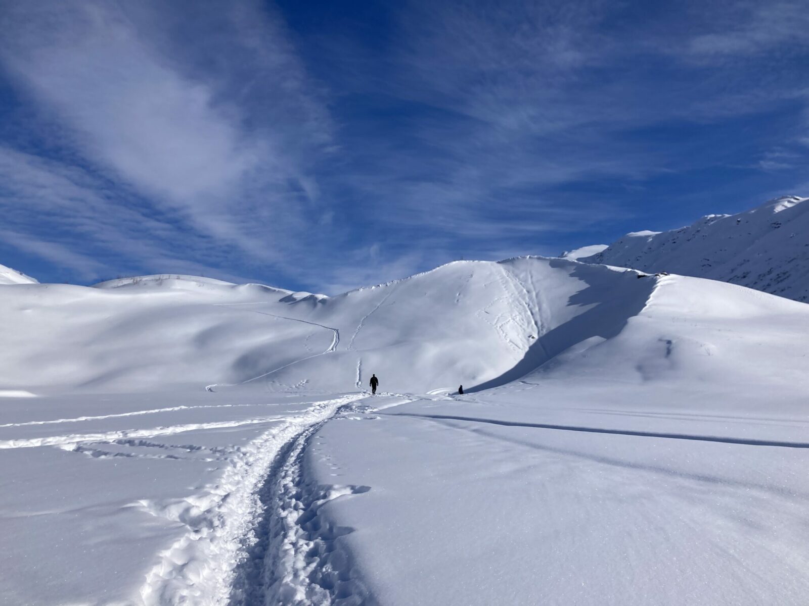 Two hikers in the distance on a packed snowy trail in a valley between mountains approaching Castner Glacier