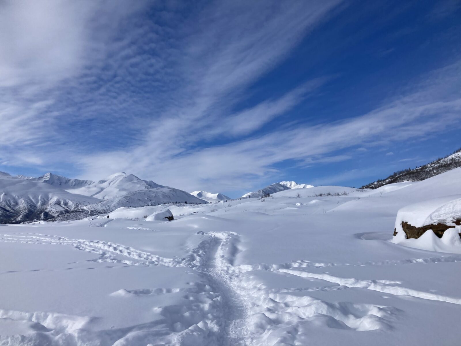 A packed trail in the snow in a valley surrounded by high snowy mountains on a mostly sunny day