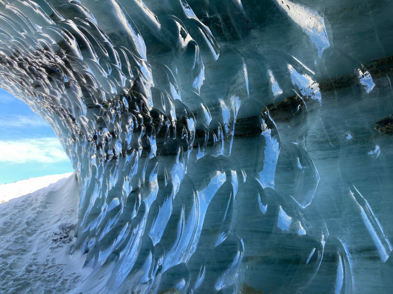 blue scallop shaped ice walls of an ice cave under Castner glacier. there are gravel brown streaks in the blue ice and the suns reflection is hitting it even though it is not in direct sunlight. There is a bit of snow around the cave entrance