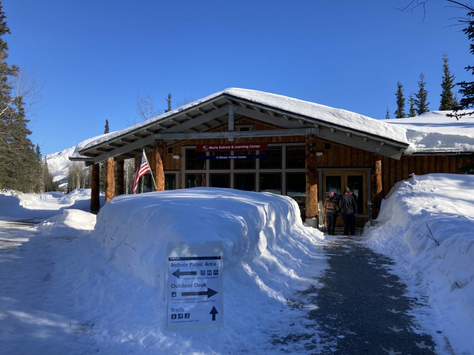 The winter visitor center in Denali National Park. It is a log building with an american flag in front and two people are approaching the door. Deep snow surrounds the building and there is a sign indicating trails and the outdoor desk and indoor picnic area