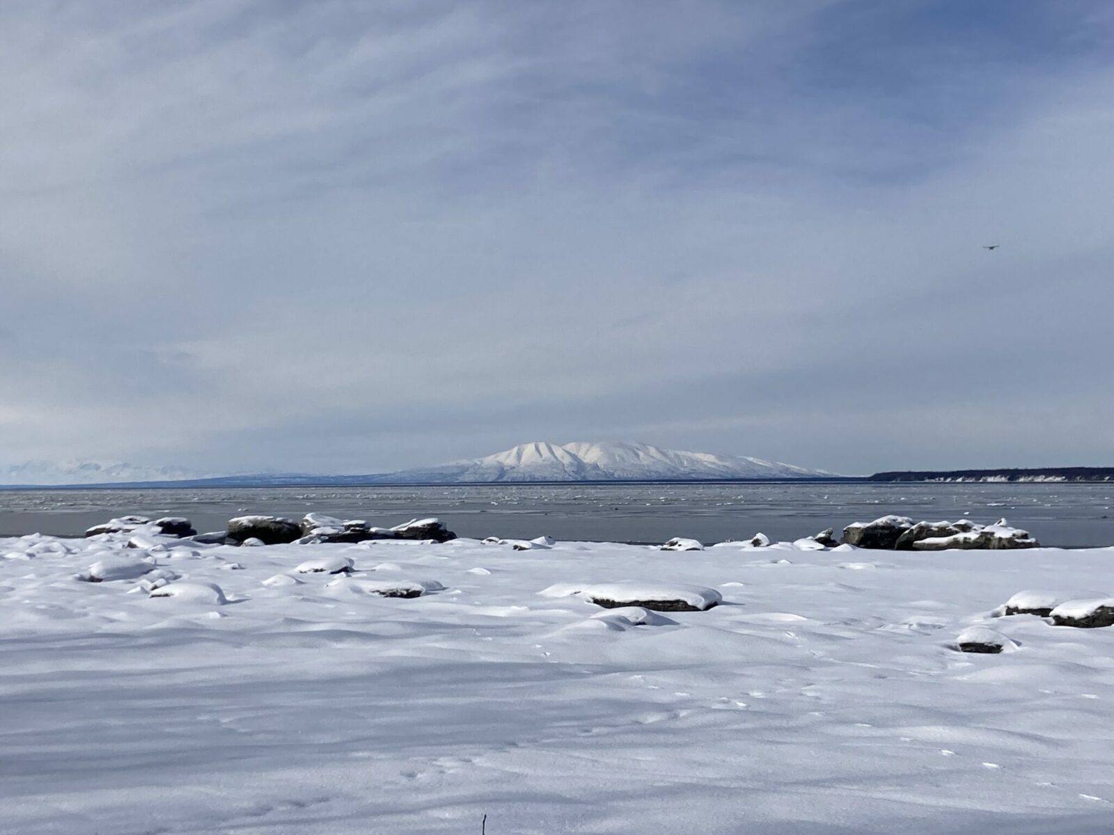 A view across a snowy rocky area and water with ice in it to a snow covered mountain in Anchorage Alaska.