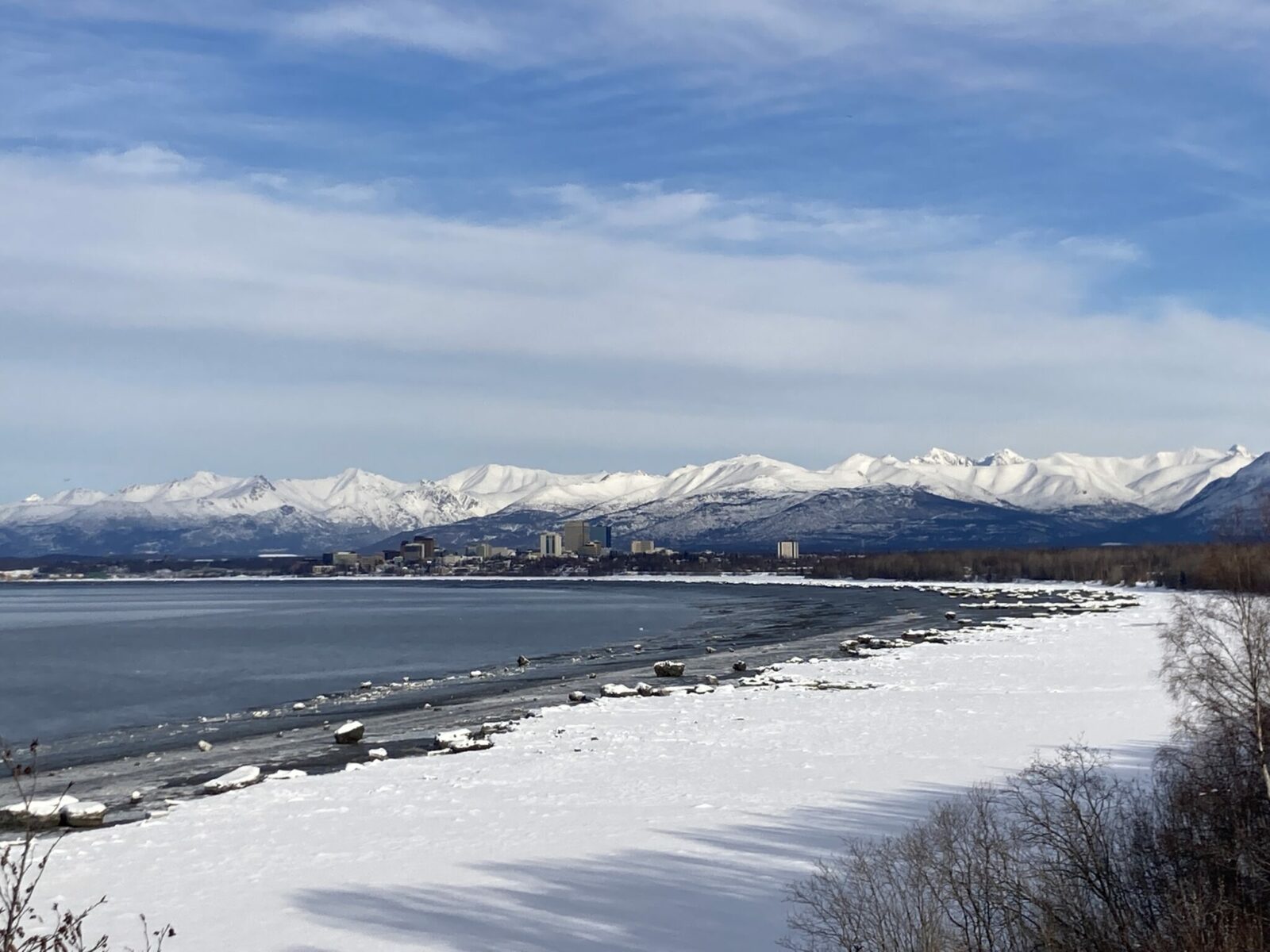 A snowy beach and water with the skyline of Anchorage Alaska and nearby mountains in the distance.