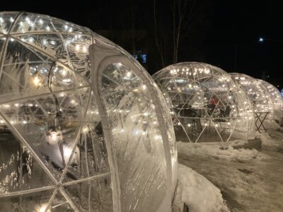 Several clear plastic tents with white lights and dining tables inside them.