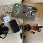 a pile of electronic devices and cables, including an apple laptop, bluetooth headphones, a go pro, a garmin inreach and several cables