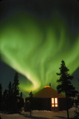 Green northern lights streaking up into the ski above a yurt with lights on inside and trees at Chena Hot Springs