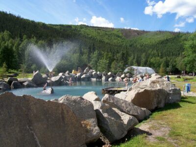 An outdoor pool surrounded by rocks and green grass and forest on a sunny summer day at Chena hot springs