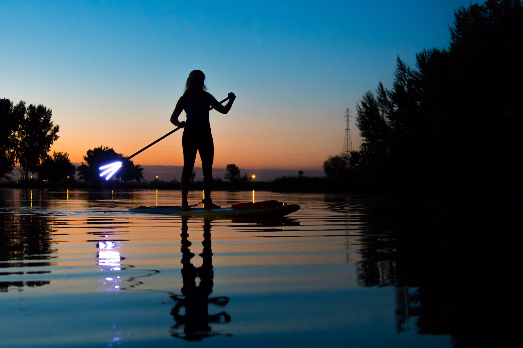 A person on a paddleboard with a lighted paddle at sunset. The person is silouetted against the sky on the river near Tri-Cities