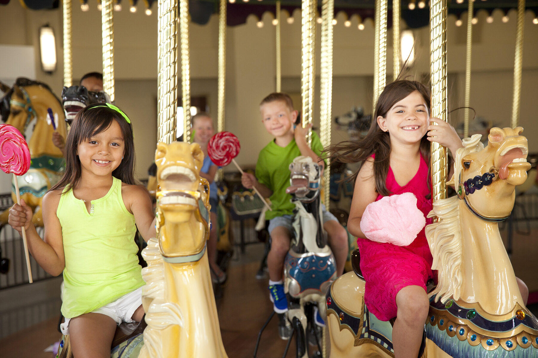 Two girls in brightly colored outfits riding horses on a historic carousel. One of the girls has a giant lollipop in her hand. There are other kids blurred in the background