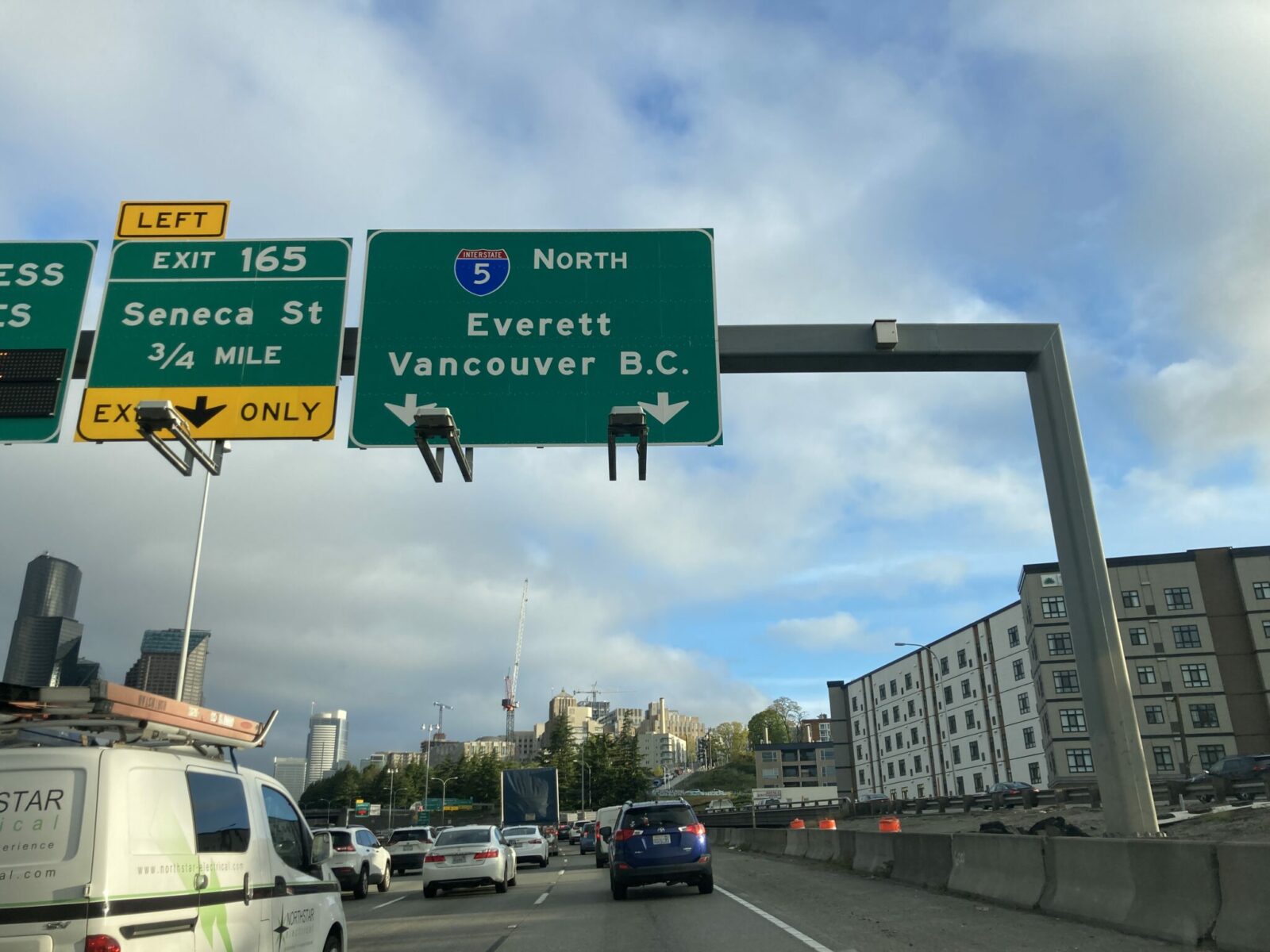 A picture taken from the passenger seat of a car on the freeway in downtown Seattle. Road signs point to a downtown exit and also I-5 north to Everett and Vancouver BC