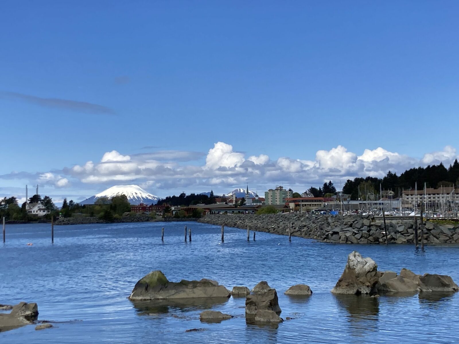 A few rocks in the water in the foreground and in the background the port of Sitka. In the distance is a snow covered volcano