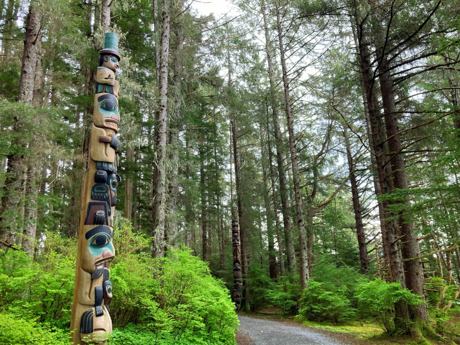 A wooden totem pole with characters painted in green, black and red next to a wide gravel trail in the forest in Sitka, Alaska